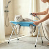 A person is using a foldable pet bathing stand to wash a fluffy gray cat under a shower head.