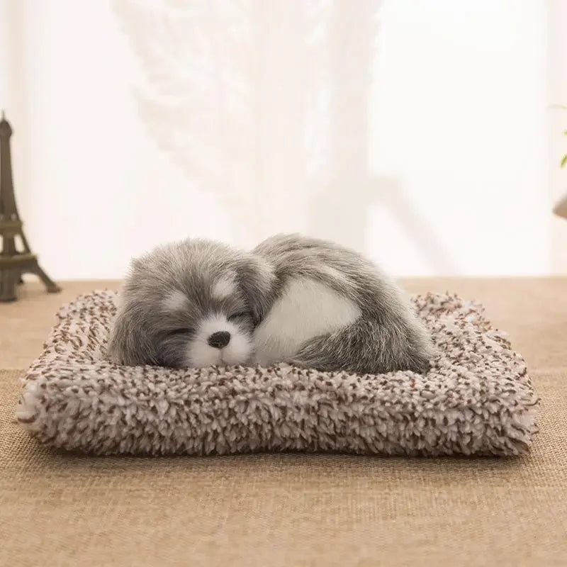 Ein kleiner, flauschiger grau-weißer Schlafhund-Plüschtier auf einer beigeen Stoffoberfläche.