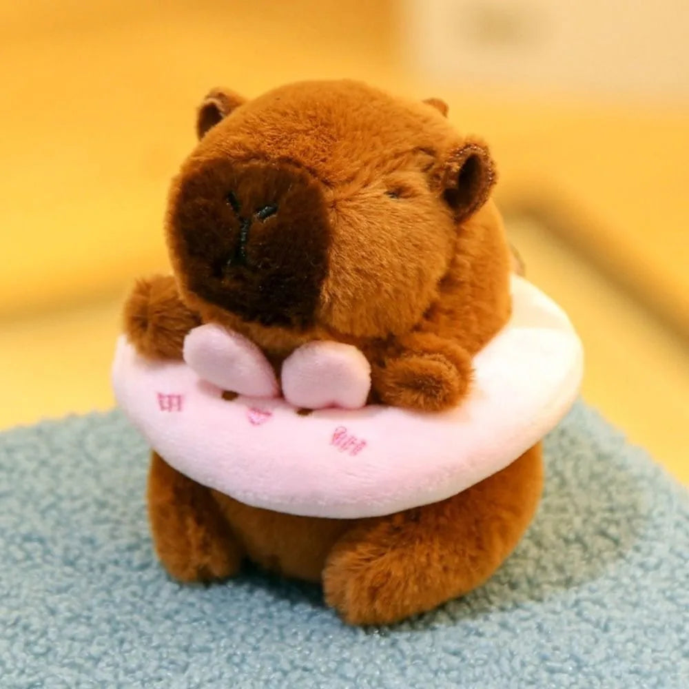 Braune Capybara-Plüschpuppe mit pinkem Hoodie und Flügel, sitzt auf blauer Oberfläche.