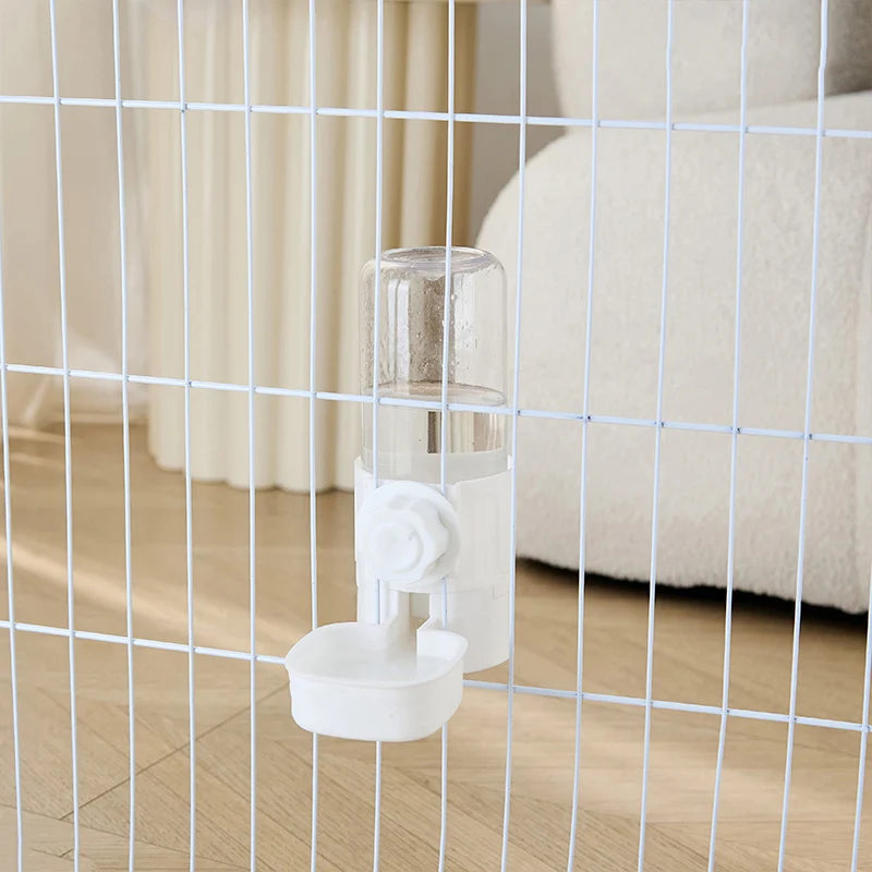 A clear automatic pet feeder waterer is mounted on a wire grid in a light-colored room.