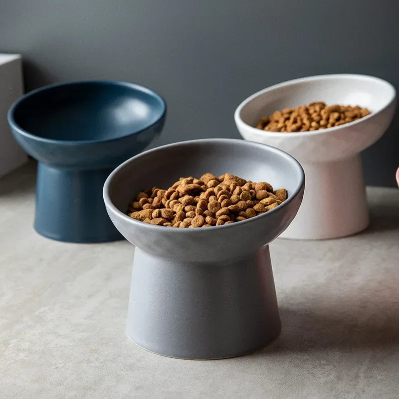 Three elevated ceramic food bowls in shades of blue, gray, and white, filled with dry pet food.