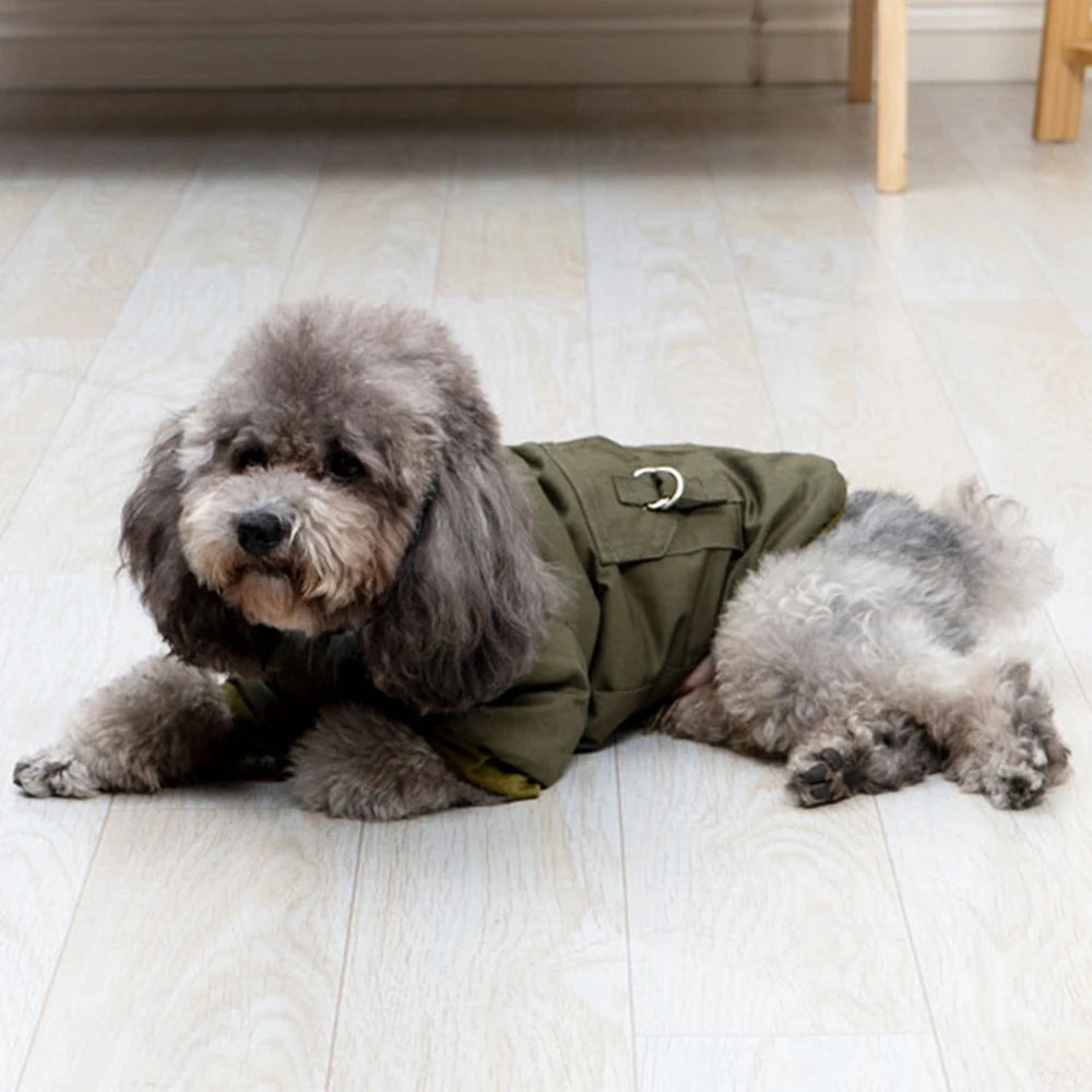 A fluffy grey poodle wearing an olive green hooded jacket lying on a light wooden floor.