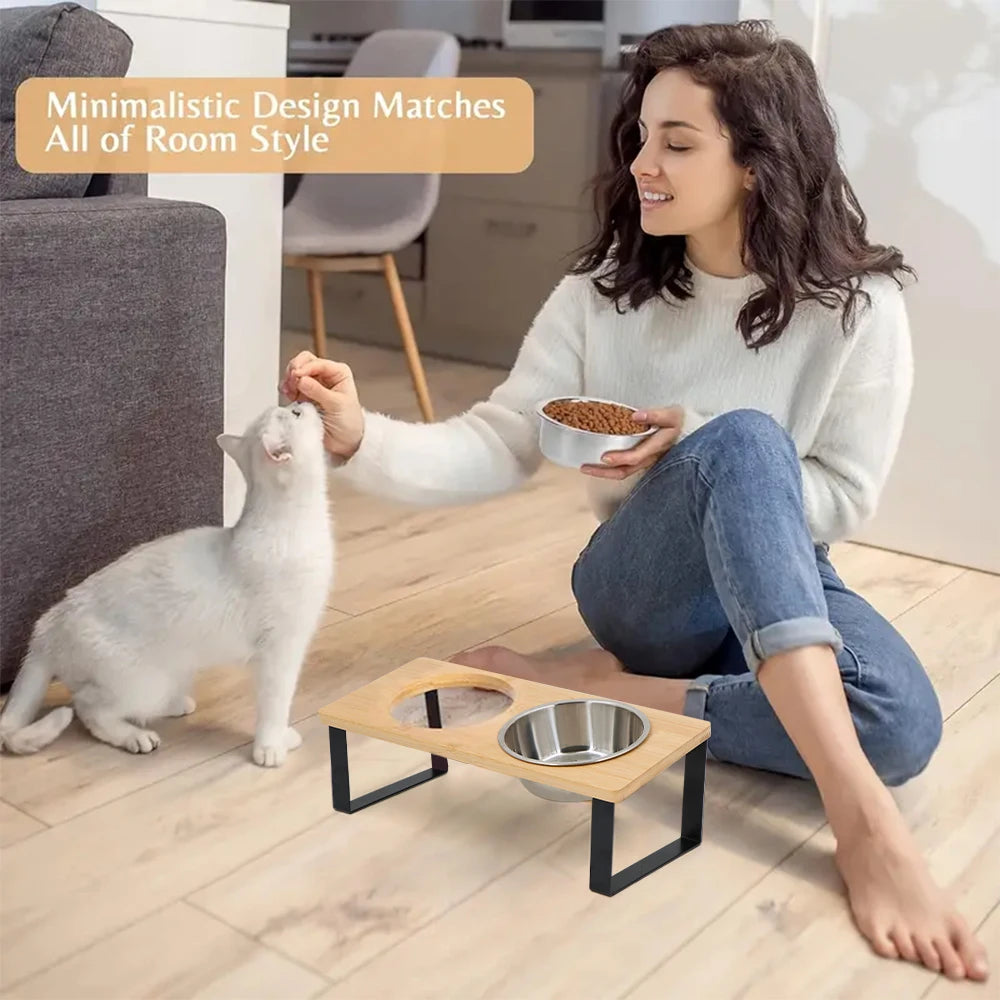 A woman is feeding a white cat from an elevated wooden pet bowl with a stainless steel feeding stand on a light wood floor.