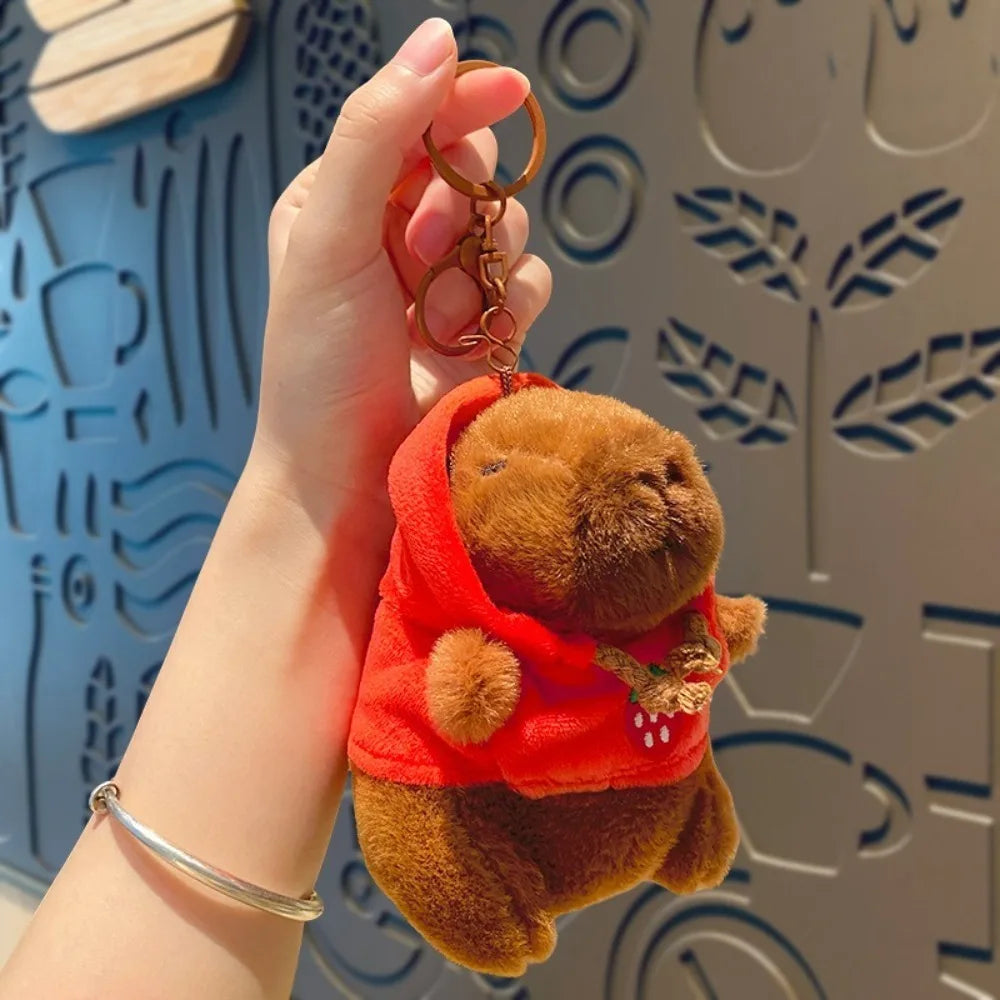 Eine Hand hält eine braune Capybara-Plüschpuppe mit orangefarbener Kapuzenjacke und goldenen Ohrringen vor einem gemusterten 
