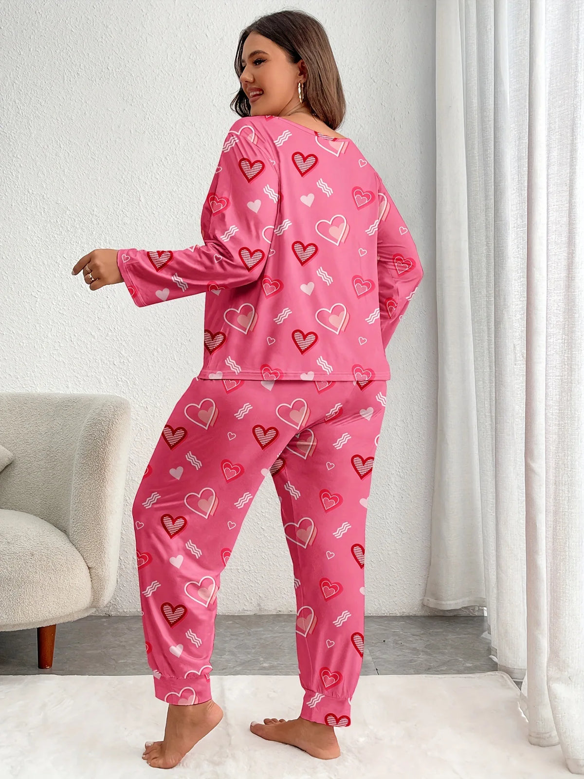 A woman models a pink heart print pajama set featuring small red hearts on a long-sleeved top and matching pants.