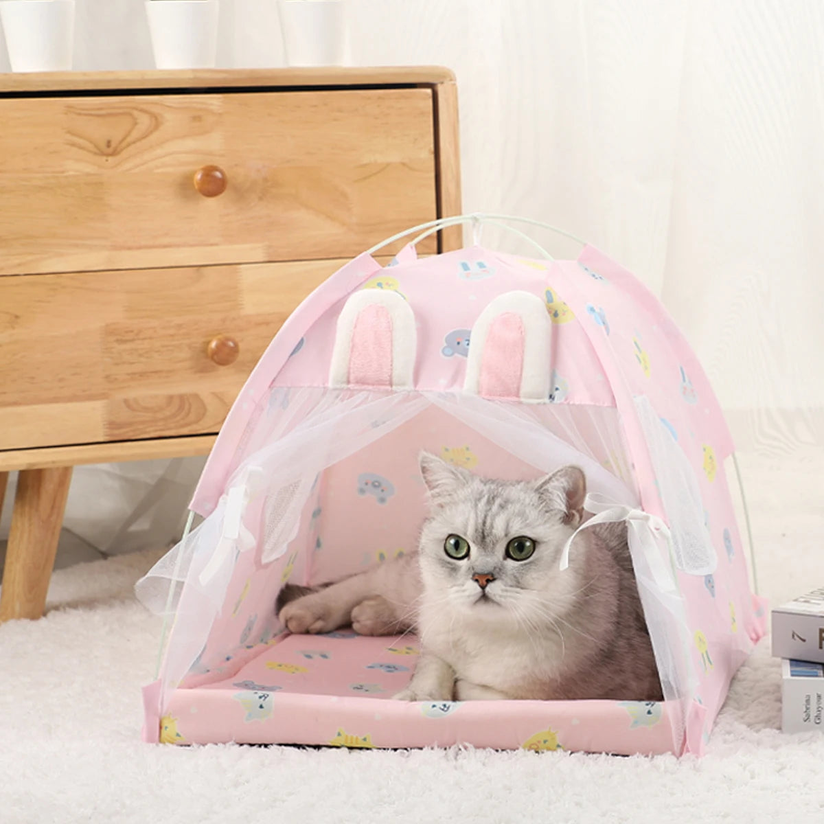 Un chat gris se repose dans un lit de tente en filet rose sur un tapis blanc à côté d'une commode en bois.