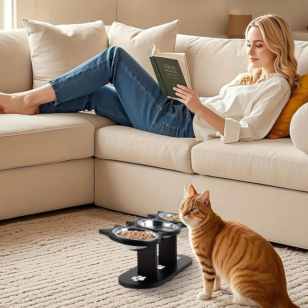 A ginger cat sits in front of a beige sofa with an open book, next to an elevated cat food bowl stand.