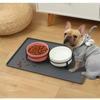 A light gray dog wearing a gray hoodie looks at a pink bowl of dry pet food on a dark feeding mat.