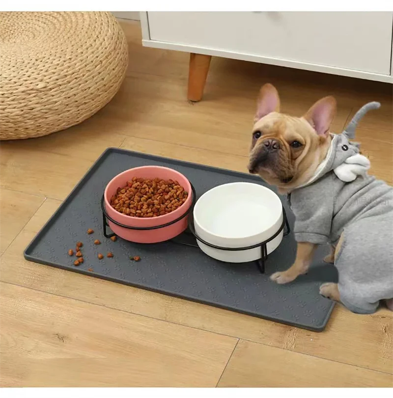 A light gray dog wearing a gray hoodie looks at a pink bowl of dry pet food on a dark feeding mat.