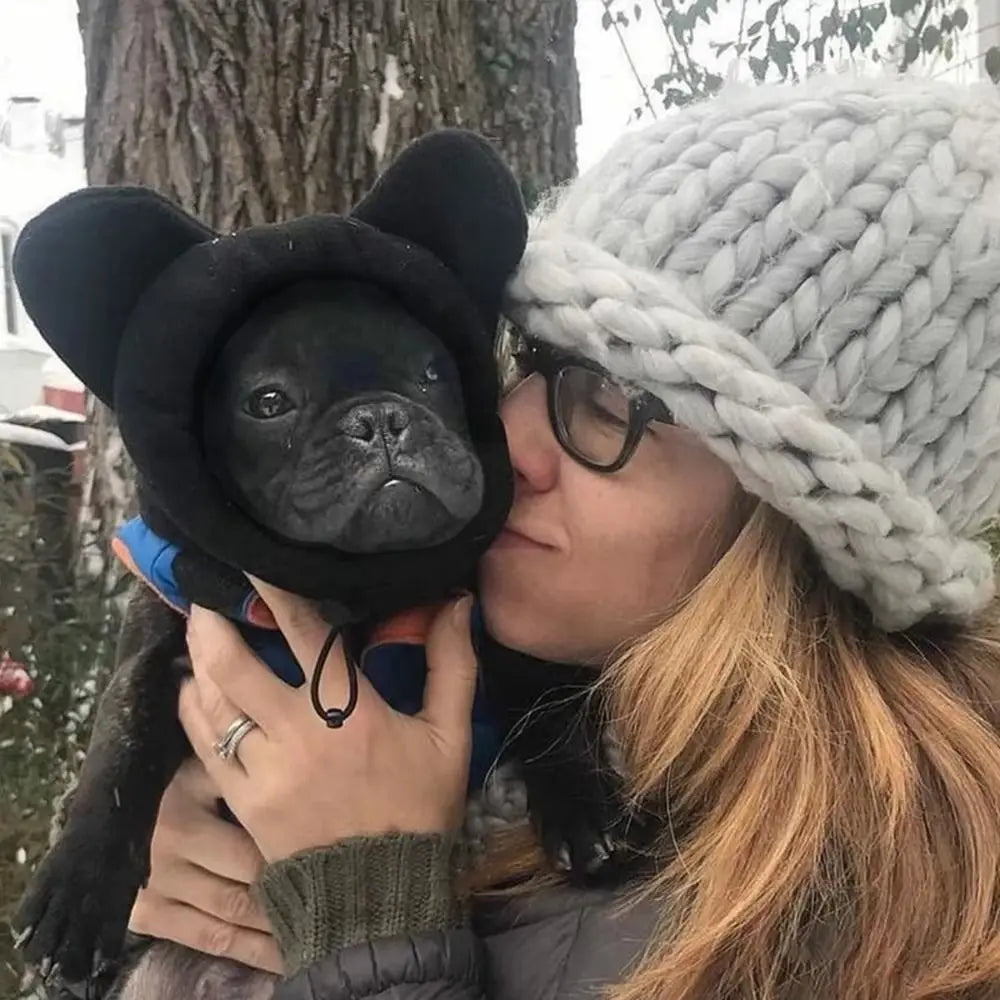 Una mujer besa a un perro negro que lleva un gorro de orejas negras mientras sostiene un gorro de invierno tejido gris claro.