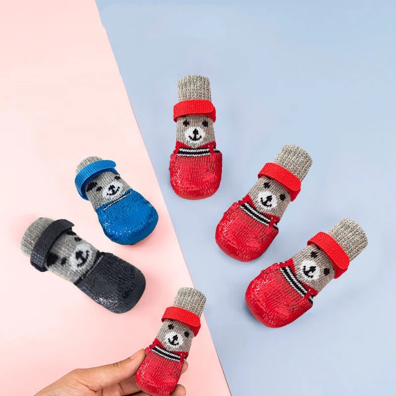 A hand holds a pair of pet paw protection socks featuring cute dog and cat faces in gray, red, and blue.