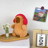 Flauschiger brauner Capybara-Plüschtier mit roher Kapuzen sitzt neben einem kleinen Glas Blumen und einem gerahmten Kunstwerk