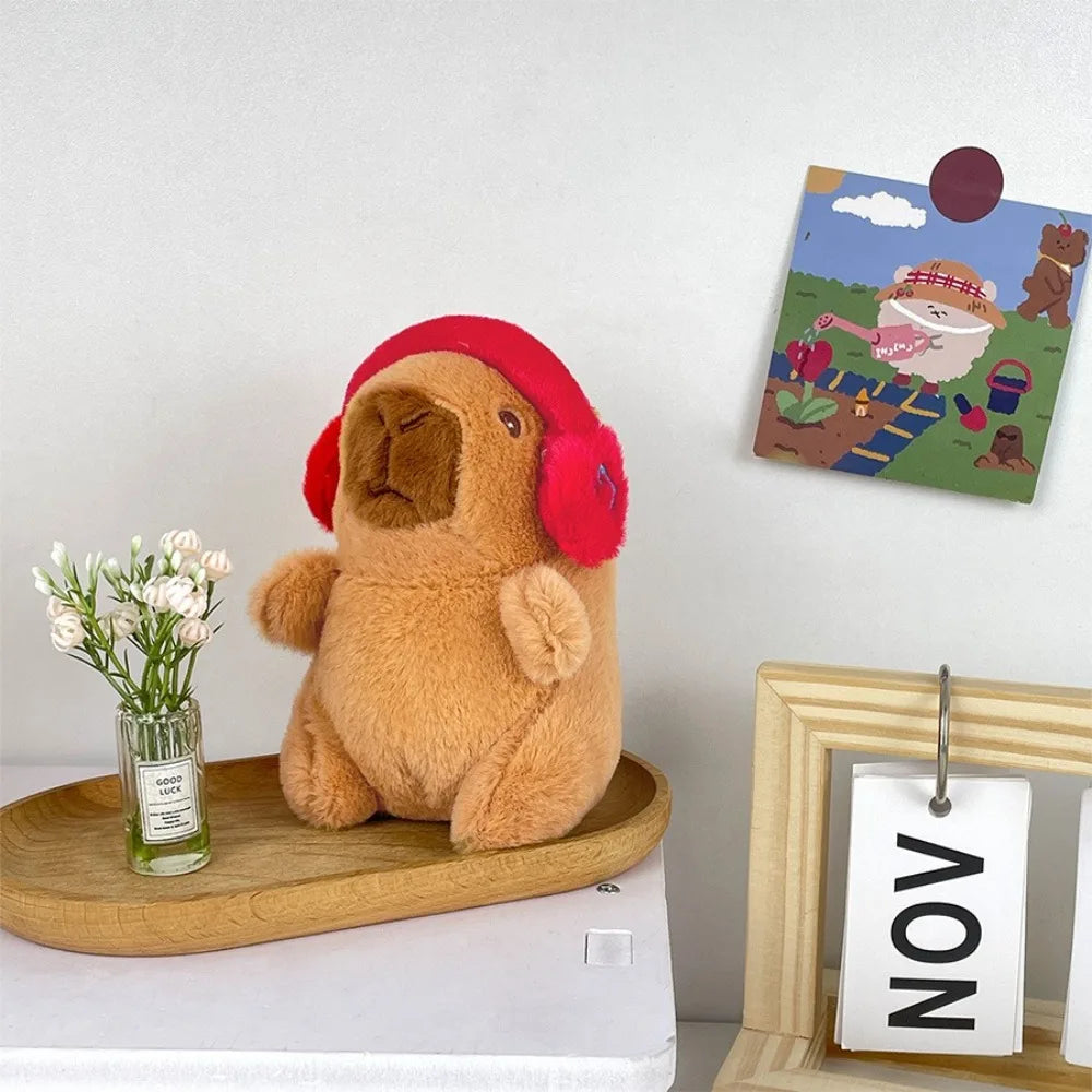 Flauschiger brauner Capybara-Plüschtier mit roher Kapuzen sitzt neben einem kleinen Glas Blumen und einem gerahmten Kunstwerk