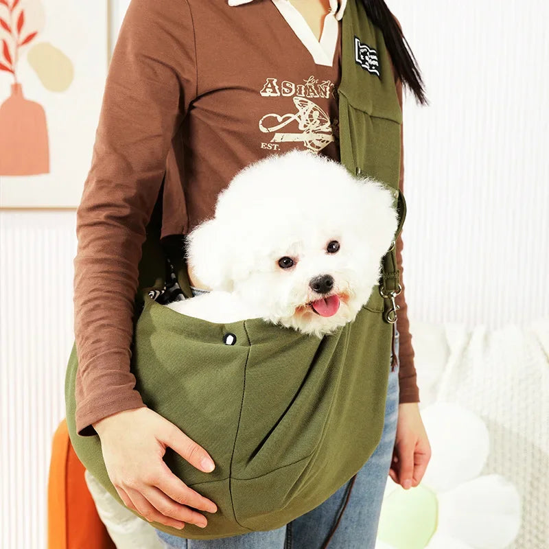 Eine Person trägt einen olivgrünen Sling-Carrier und hält einen flauschigen weißen Hund vor sich.