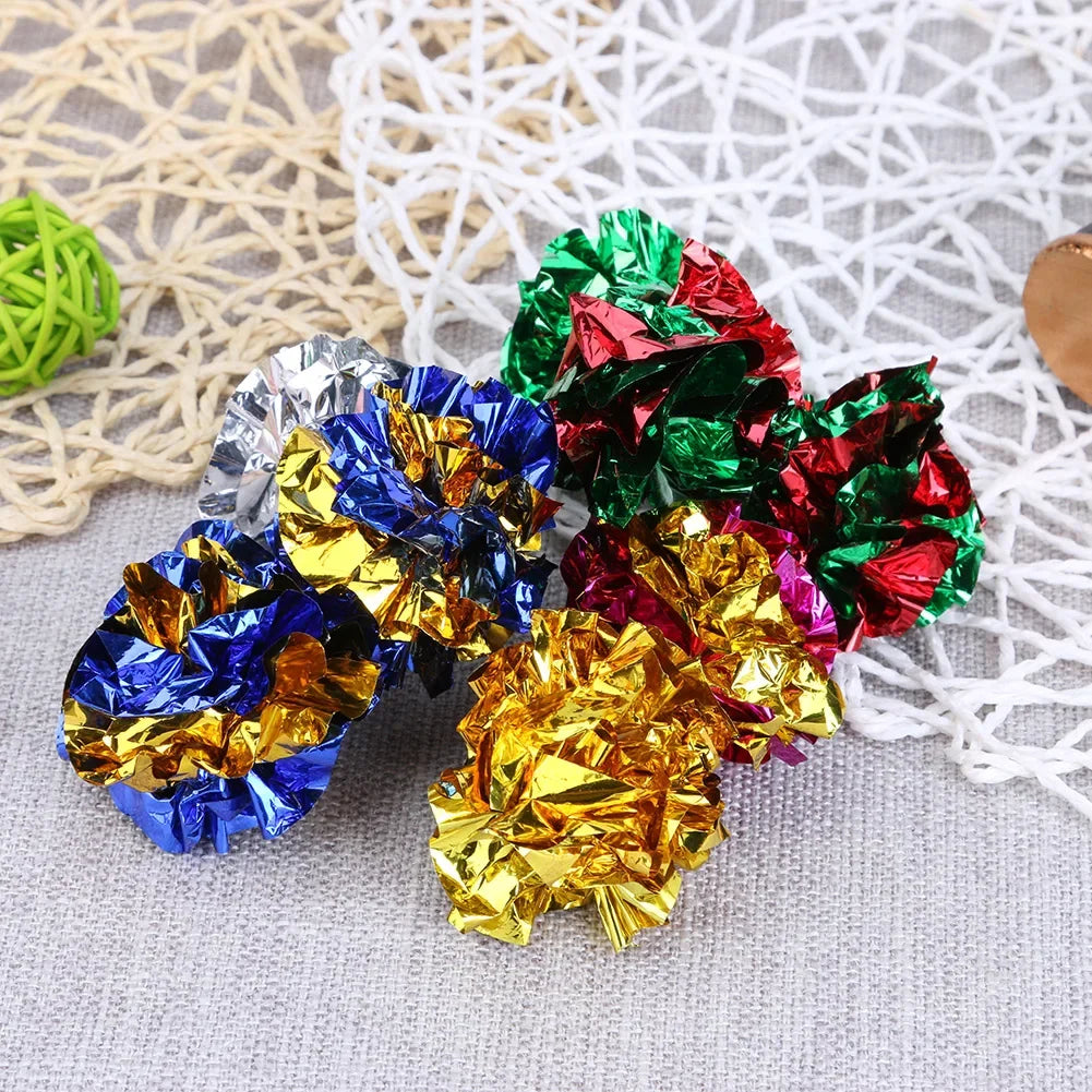 A collection of colorful crinkle balls in green, red, blue, and gold are displayed on a light gray fabric.