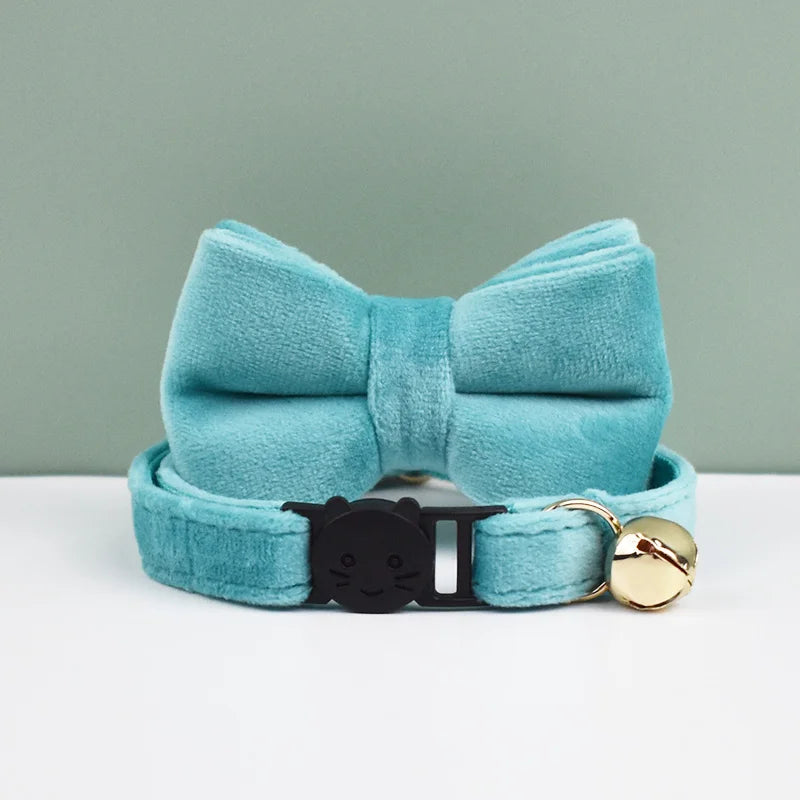 A teal velvet cat collar with a bow knot and a gold safety buckle resting on a white surface.