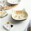 A ceramic cat-shaped sauce dish with orange stripes rests on a white cloth next to a natural fiber mat.