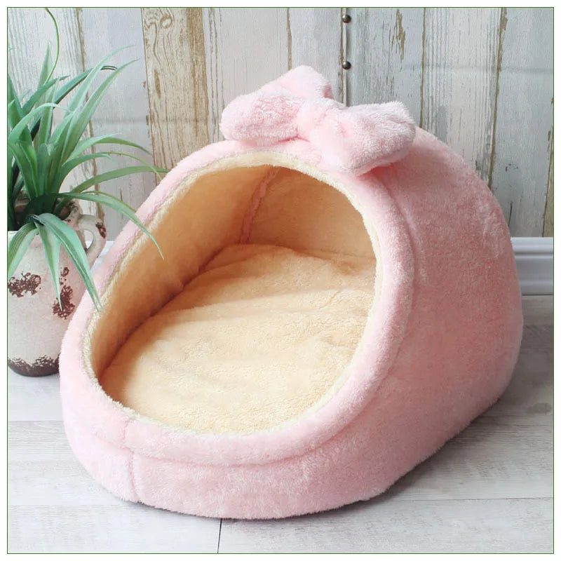 A pink, fluffy pet bed shaped like a cave with a bow, resting on a light wooden floor.