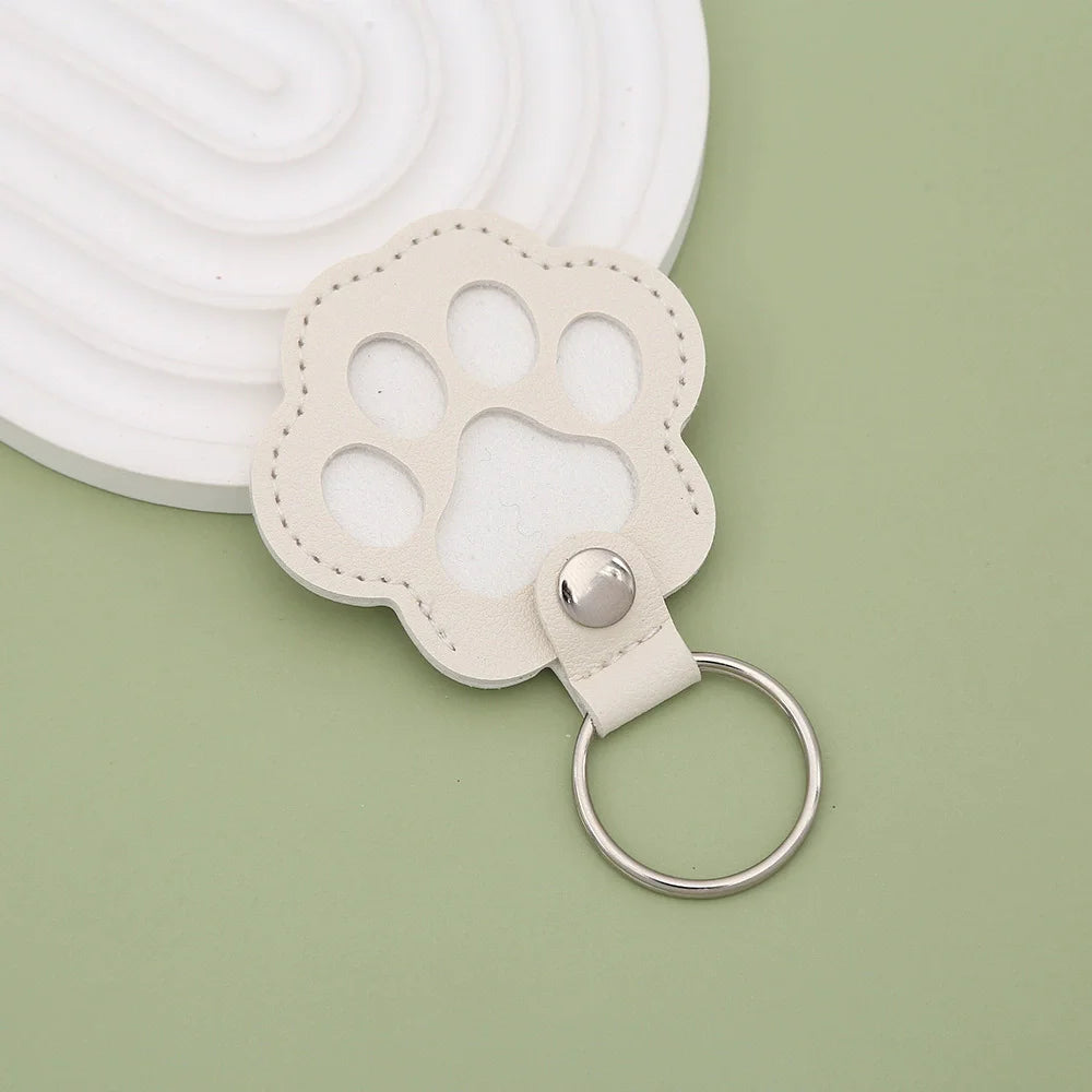 A light beige leather pet paw print keepsake with a metal ring attached, set against a pale green background.