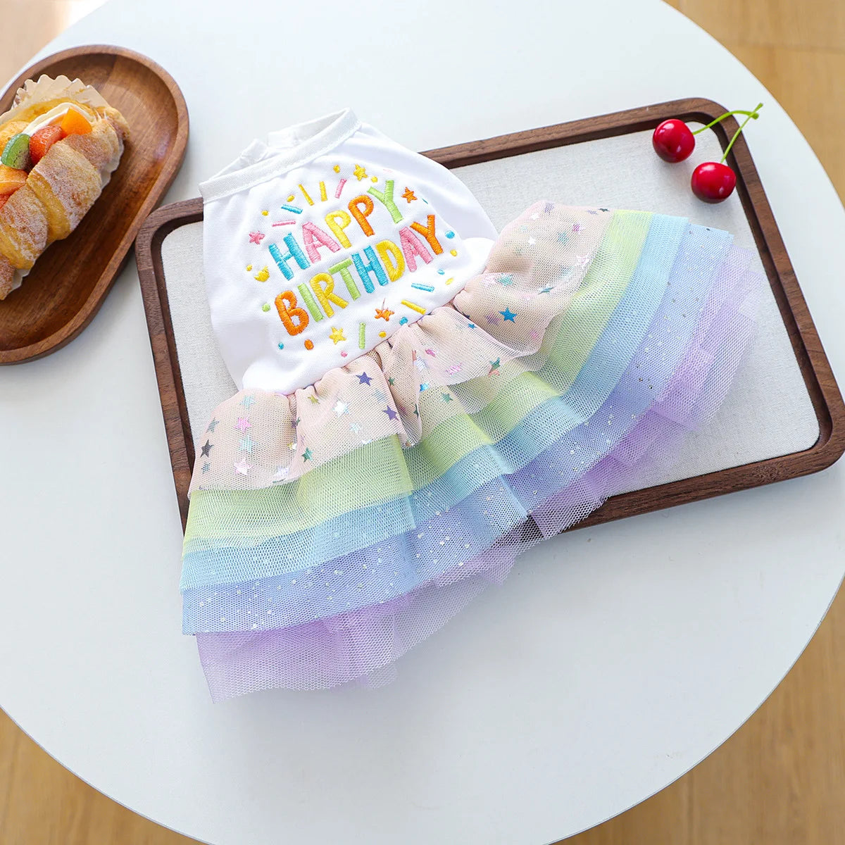 A white dog birthday suit with colorful tulle layers is displayed on a wooden tray next to a pastry.