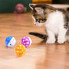 A kitten is sitting next to several colorful plastic toy balls shaped like jingle bells on a wooden floor.