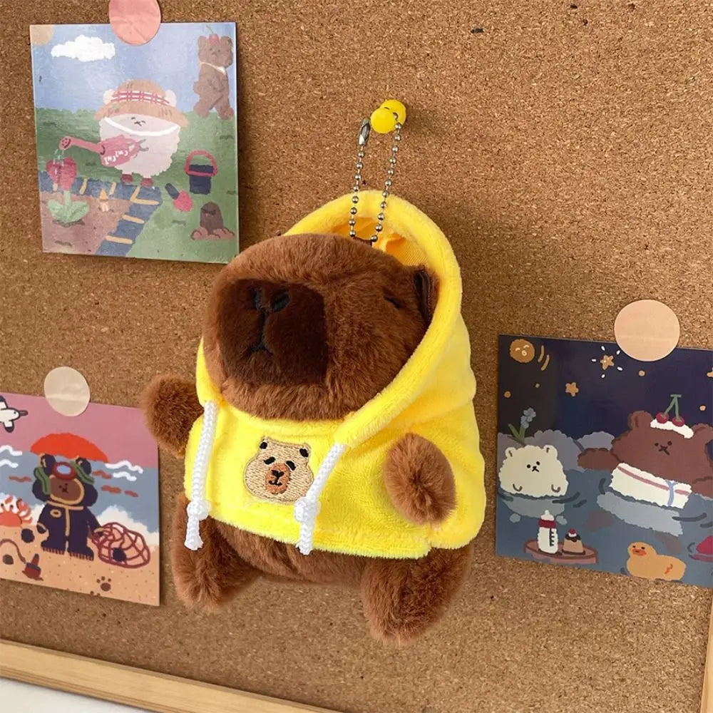 Braune Capybara-Plüschpuppe mit gelbem Hoodie und weißem Rand vor einer Korkwand.