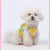 A fluffy white dog wearing a yellow mesh harness with pink floral accents.