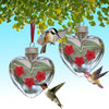 A glass heart-shaped hanging feeder with a hummingbird perched on a green leaf.