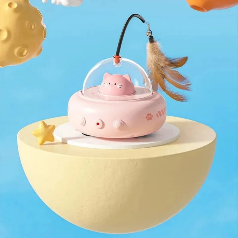 A pink automatic UFO cat teaser toy with a cat head and a feather wand is displayed on a yellow platform against a blue backg