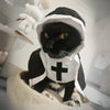 A black cat dressed in a white nun costume with a cross on the forehead, sitting on a light-colored surface.