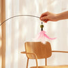 A hand is holding a feather teaser from a rotating toy, demonstrating its use near a wooden chair.