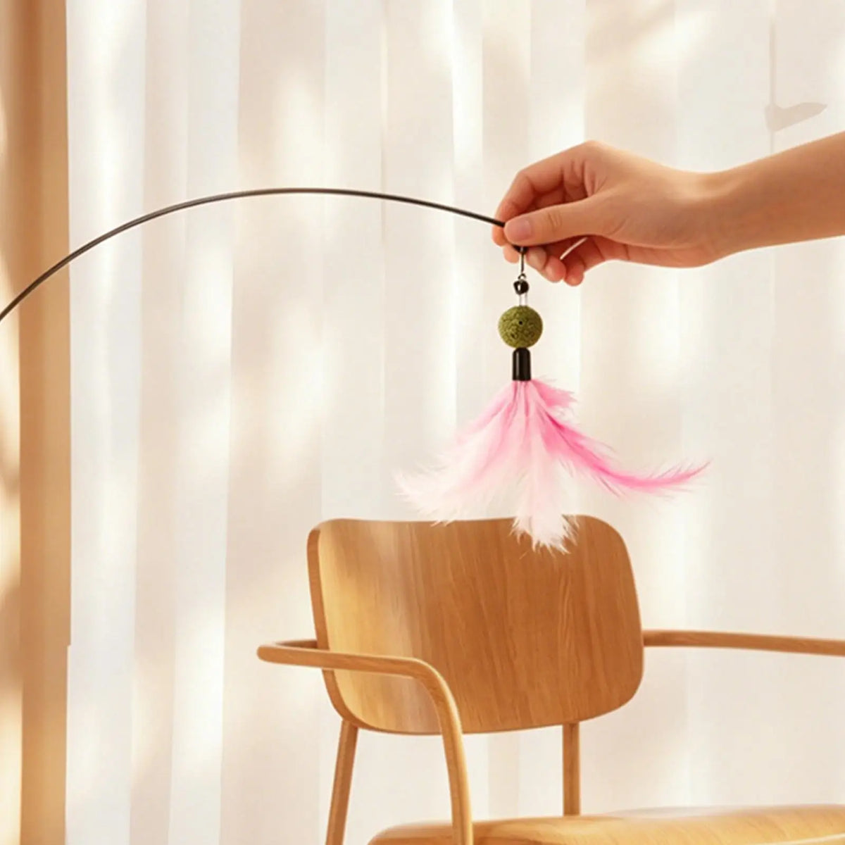 A hand is holding a feather teaser from a rotating toy, demonstrating its use near a wooden chair.