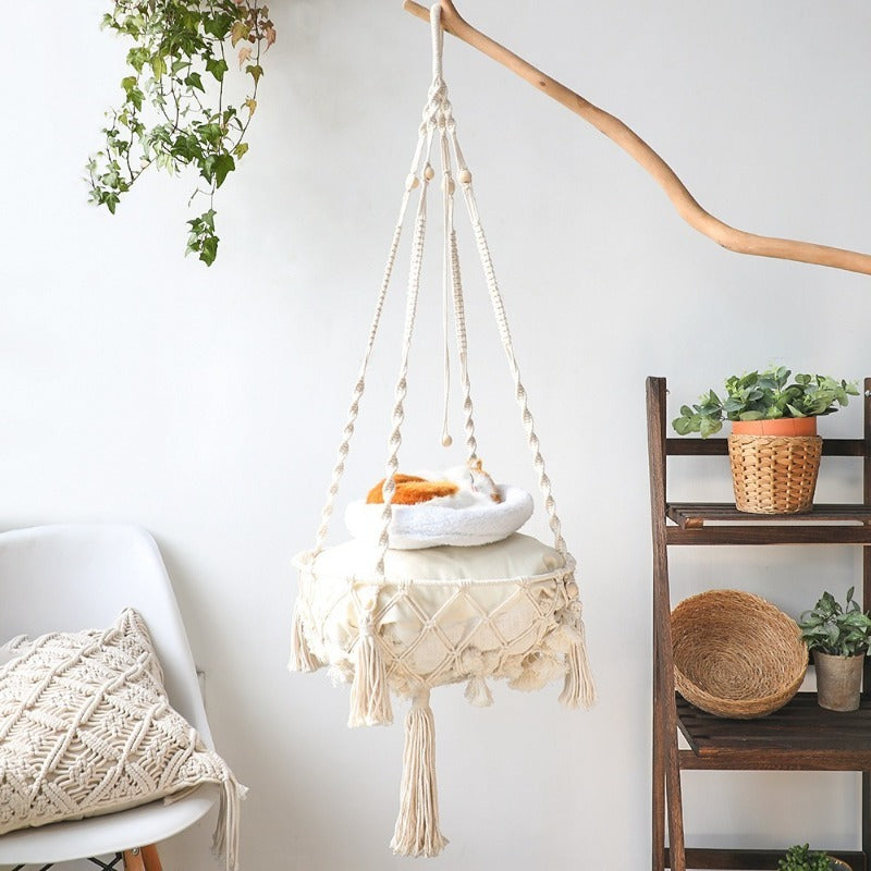 A hand-woven cotton cat hammock with a cushion is suspended by rope against a white wall.
