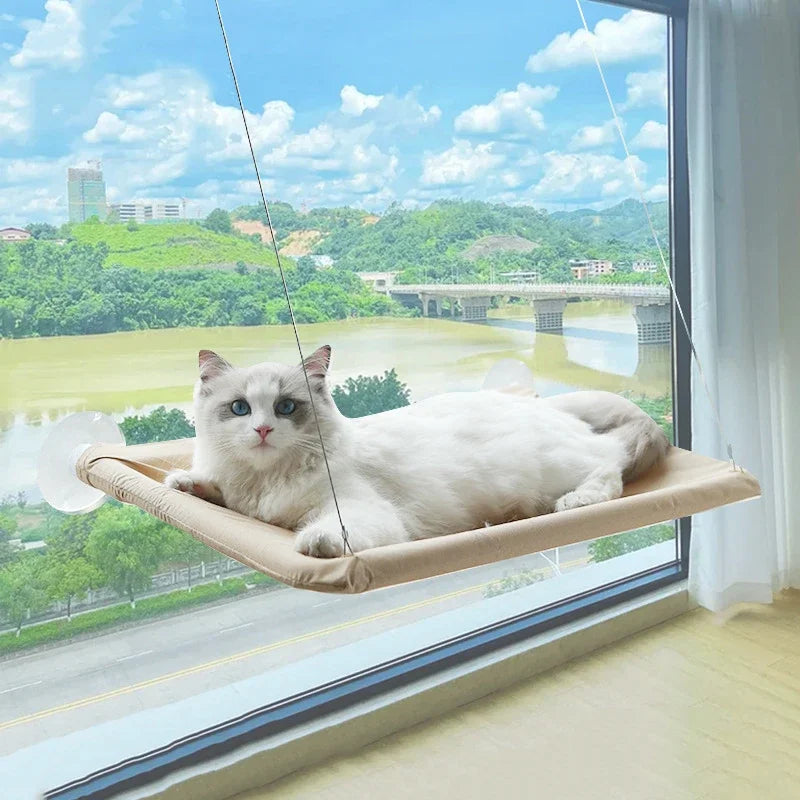 A white cat is relaxing on a beige window hammock overlooking a green landscape and distant bridge.