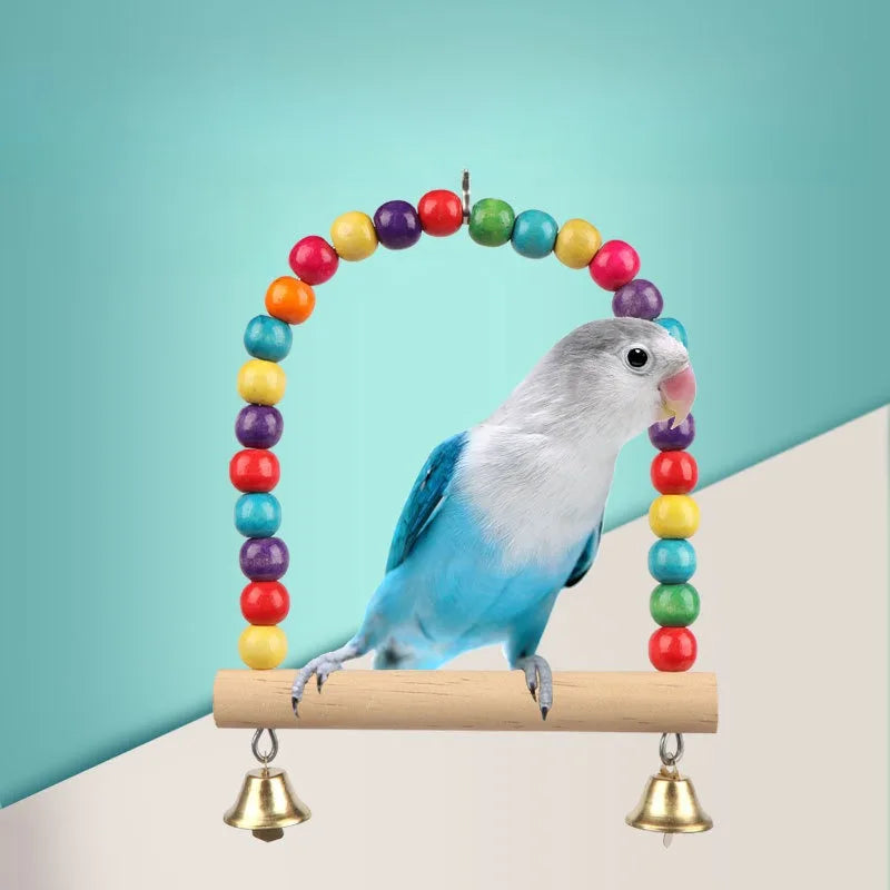 A blue and gray bird is perched on a wooden swing decorated with a colorful bead garland and suspended by brass bells.