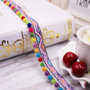 A close-up of a colorful pearl pom pom fringe lace trim draped across a white book and next to red cherries.