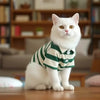A white cat wearing a green and white striped polo shirt sitting on a wooden surface.