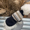 A black plaid knit sweater for dogs with white fluffy trim, resting against dried natural grasses.