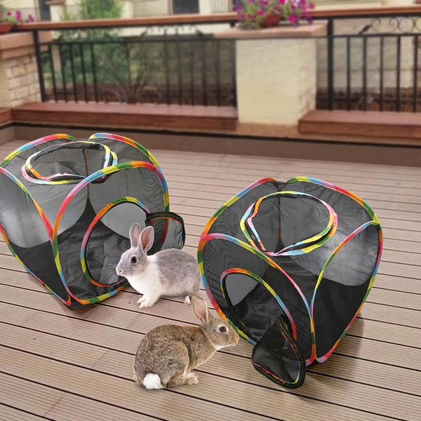 A gray rabbit sits next to a black mesh cat play tunnel with a tent and cube on a wooden deck.