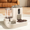 A white gravity pet food and water dispenser sits on a table, featuring a transparent water reservoir and a compartment for c
