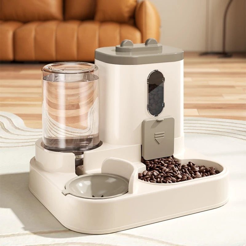 A white gravity pet food and water dispenser sits on a table, featuring a transparent water reservoir and a compartment for c