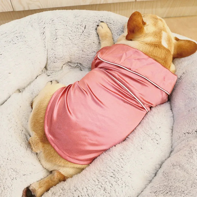 A light brown dog is lying on a white fluffy surface wearing pink silk pet pajamas.