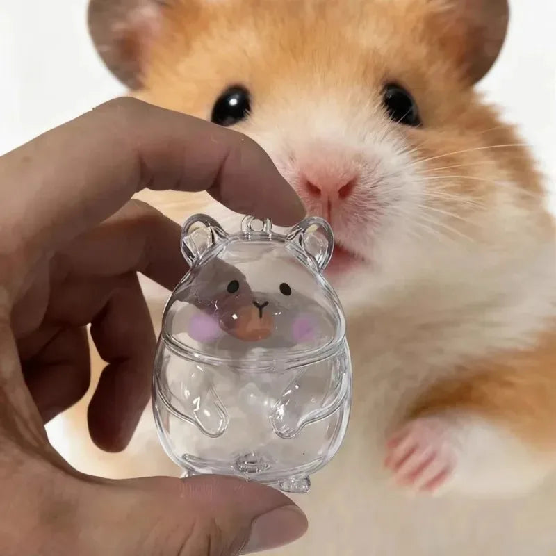 A hand holds a clear glass hamster figurine keychain in front of a fluffy orange and white hamster.