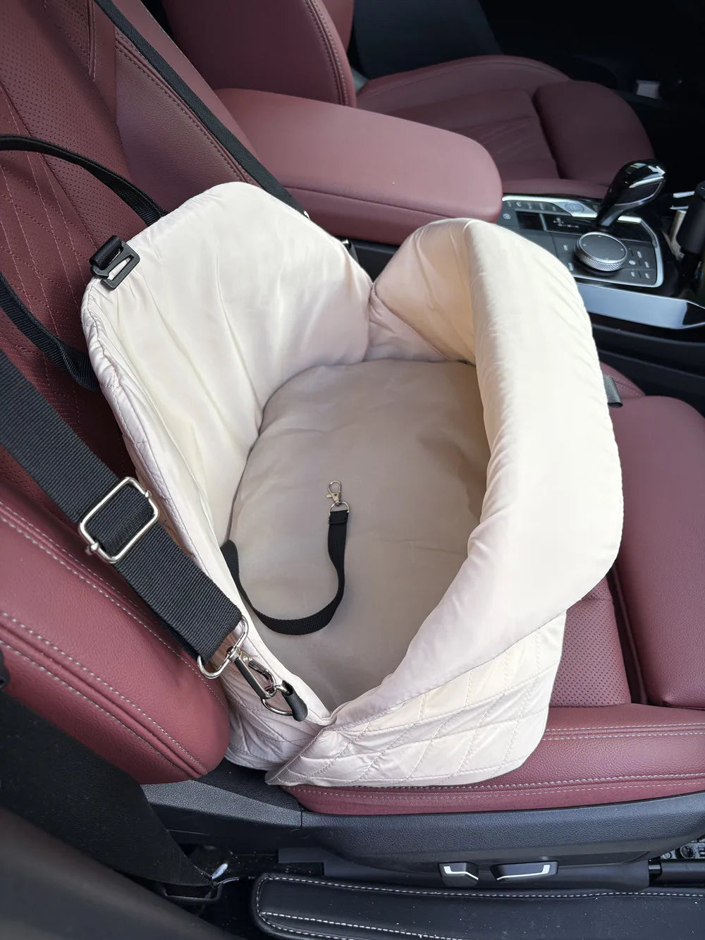 A light beige portable soft-sided pet carrier is shown placed in the seat of a car.