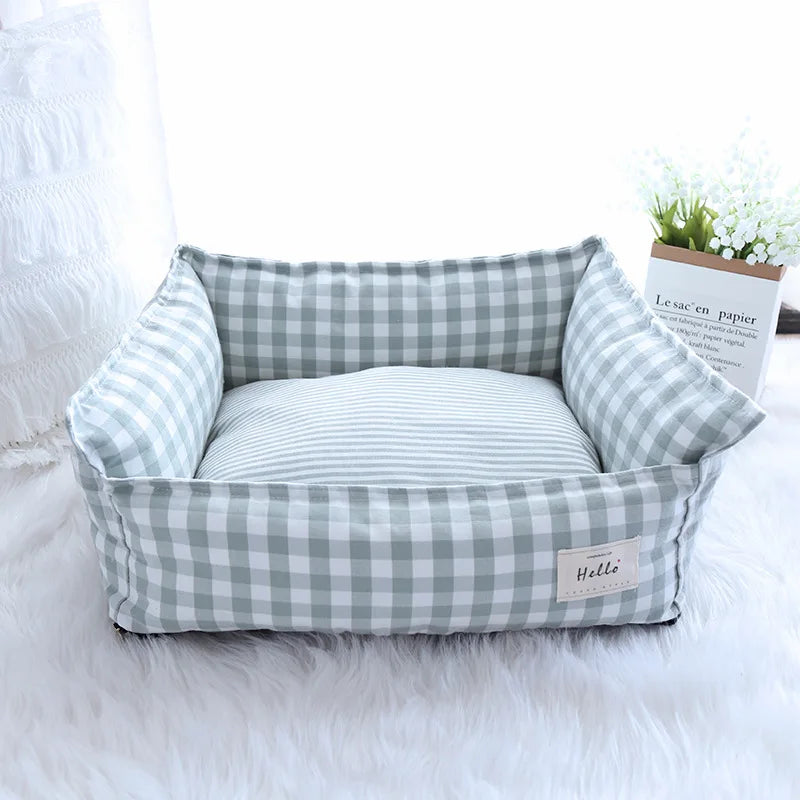 Un lit sofa pour animaux carré, rembourré, à motif vichy gris et blanc, posé sur un tapis blanc moelleux.