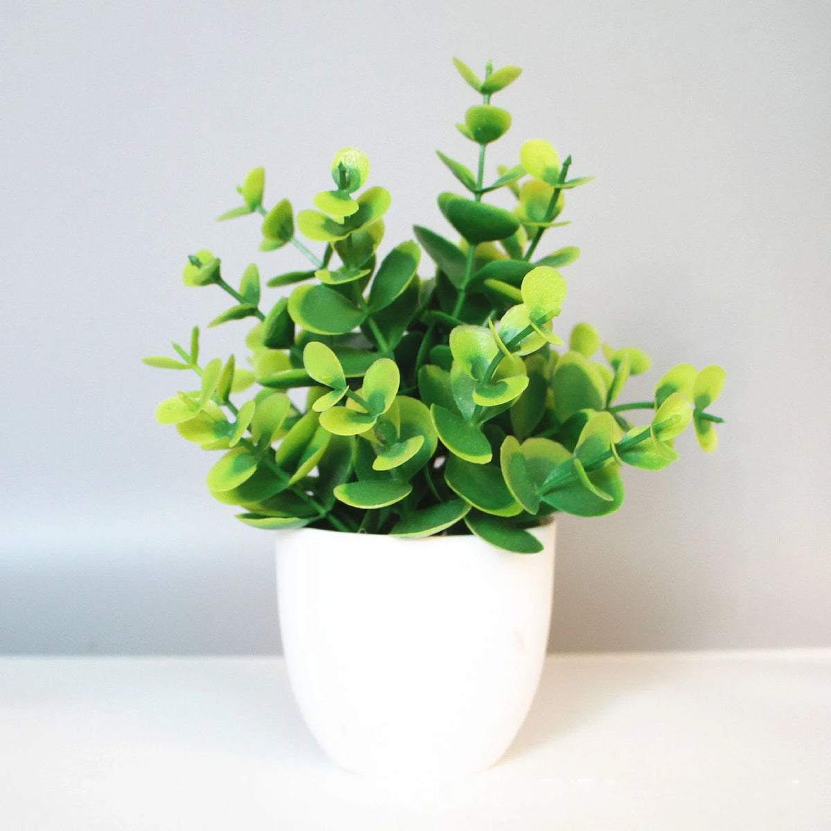 A potted artificial bonsai plant with bright green leaves sits on a white surface.