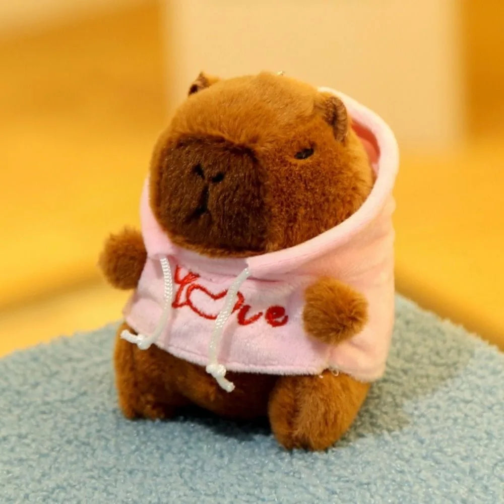 Braune Capybara-Plüschpuppe mit rosa Hoodie und roter Stickerei, sitzt auf blauer Oberfläche.