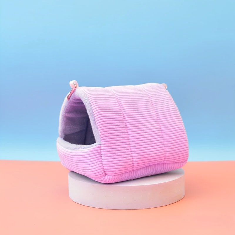 A pink, quilted cotton bird hammock nest is displayed on a light wooden surface against a soft blue background.