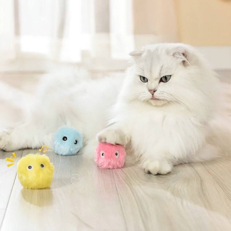 Ein flauschiger weißer Kater schaut auf bunte Plüschballen auf einer hellen Holzoberfläche.