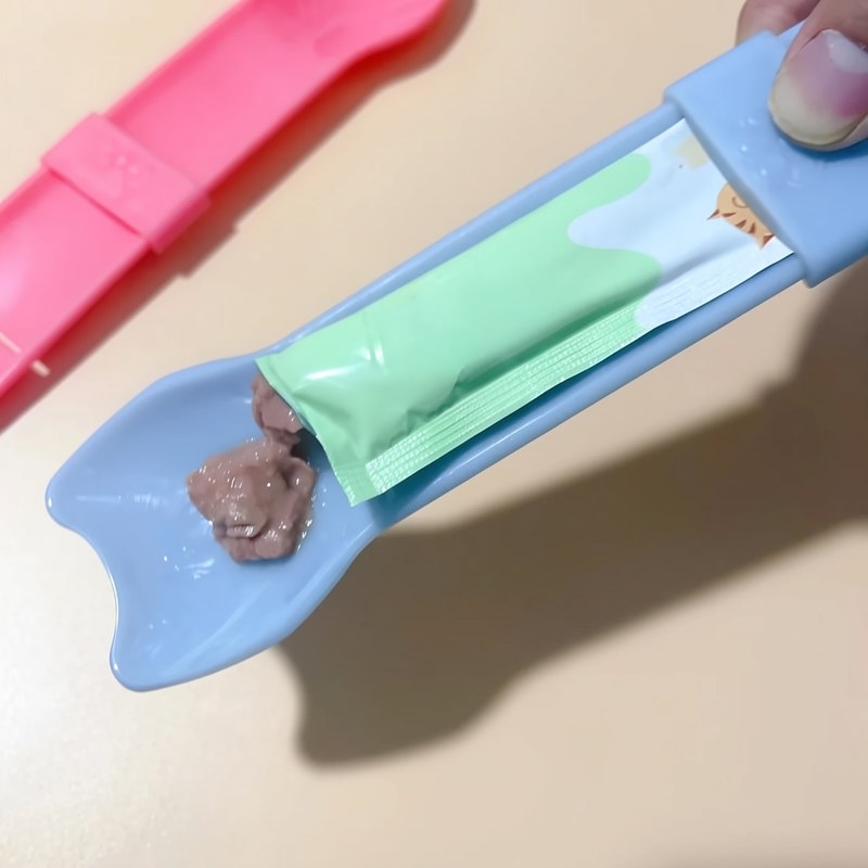 A light blue spoon is holding a green cat treat pouch, with a pink plastic tool visible in the background.